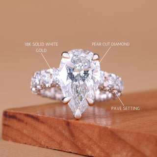 Diamond ring with labels indicating '18K Solid White Gold', 'Pear Cut Diamond', and 'Pave Setting' on a wooden surface.
